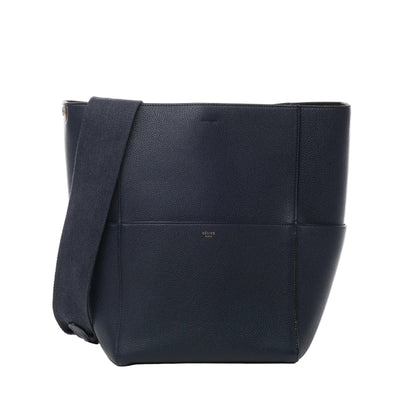 Front view of CELINE Seau Sangle Bucket Leather Shoulder Bag in navy blue, featuring pebbled leather texture, wide adjustable fabric shoulder strap, and subtle gold CELINE Paris logo against a white background.