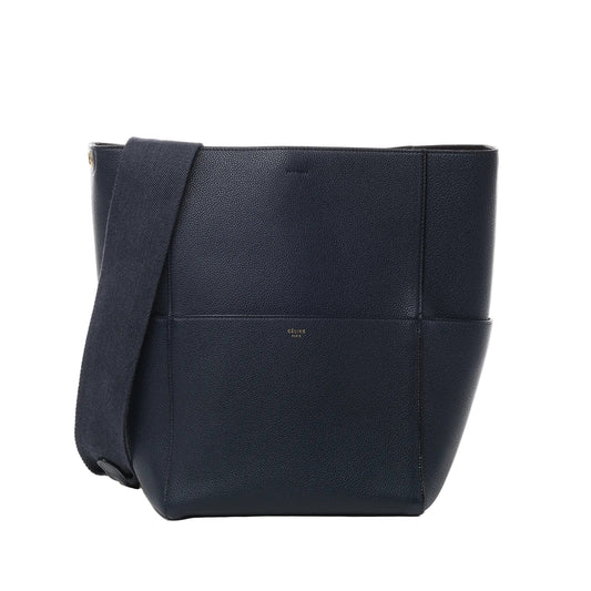 Front view of CELINE Seau Sangle Bucket Leather Shoulder Bag in navy blue, featuring pebbled leather texture, wide adjustable fabric shoulder strap, and subtle gold CELINE Paris logo against a white background.
