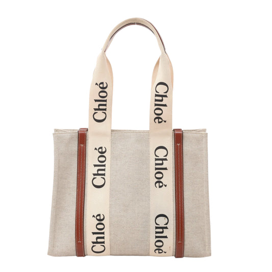 Front view of Chloé Woody Medium Tote Bag in beige canvas with brown leather trim and wide cream-colored logo webbing shoulder straps, displayed against a white background.