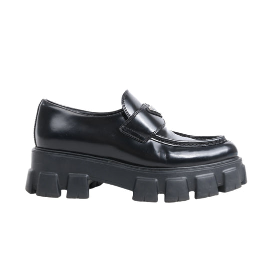 Pair of PRADA Spazzolato black leather Monolith loafers with pointy toe, chunky 55mm rubber sole, and signature silver triangle logo plaque on the strap, size 38.5 EU, photographed on a white background.
